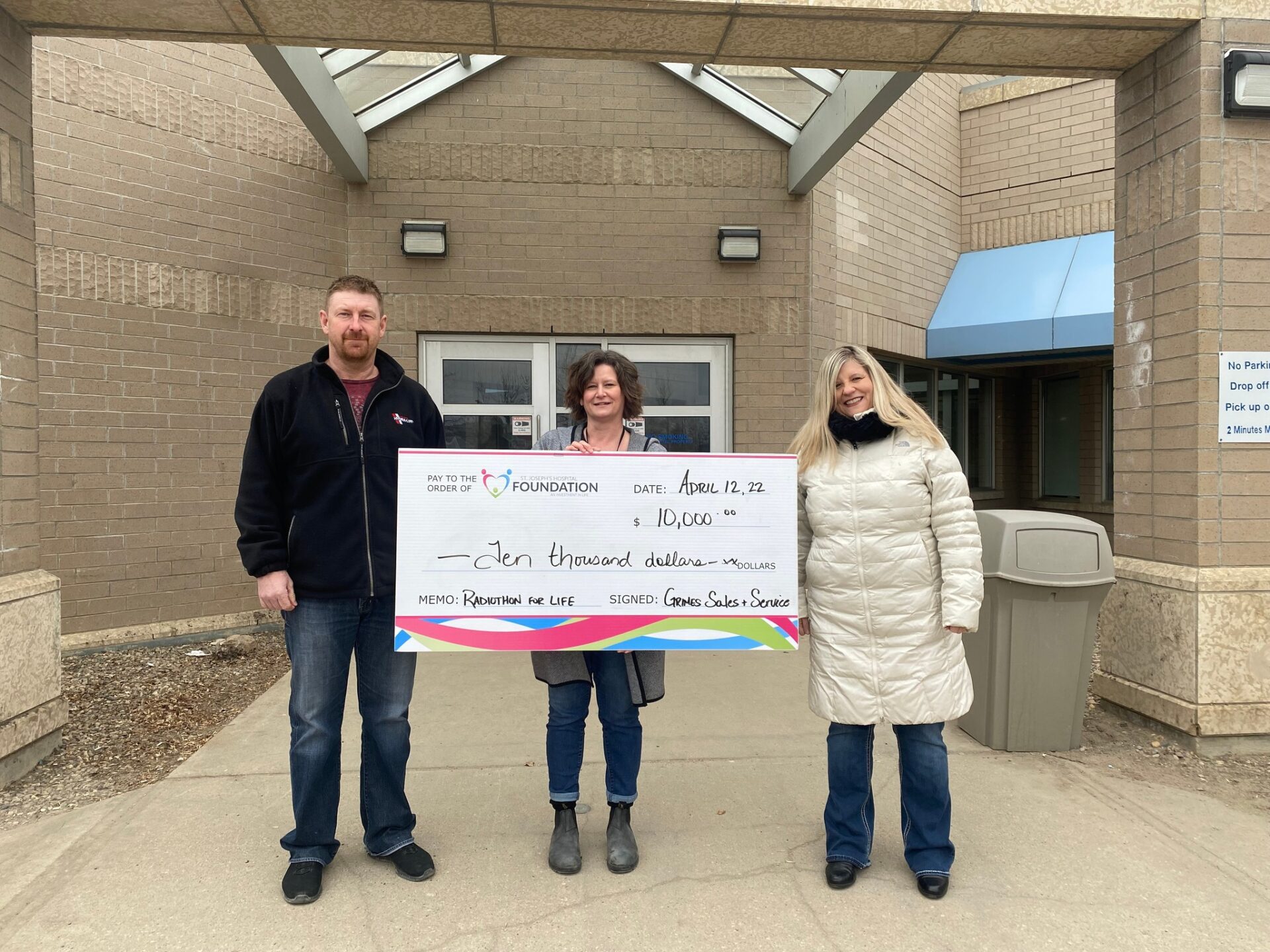 Three individuals stand in front of a building, proudly holding a large check together.