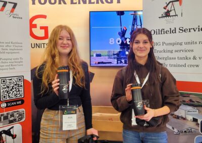 Two women stand beside a booth displaying various oil and gas signs, engaged in conversation and networking.