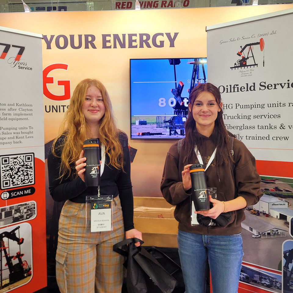 Two women stand beside a booth displaying various oil and gas signs, engaged in conversation and networking.